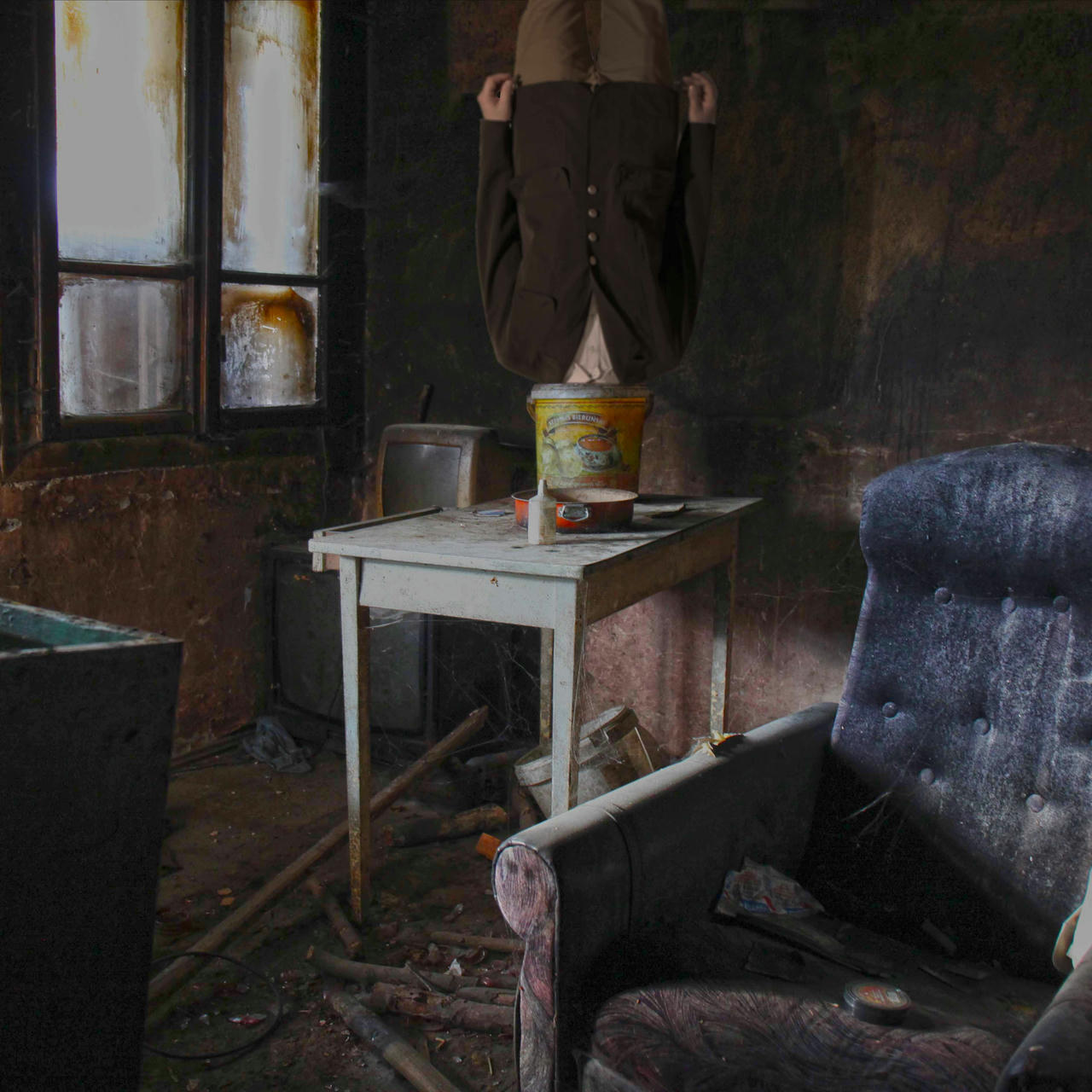 Inside a very messy, abandonned room. A white table is in the middle. There is a pan and a bucket on the table. A man is upside-down on the table, his head inside the bucket. In the back, a window with crusty opaque window panes. Old dusty TVs. In the front, a dirty blue sofa.
